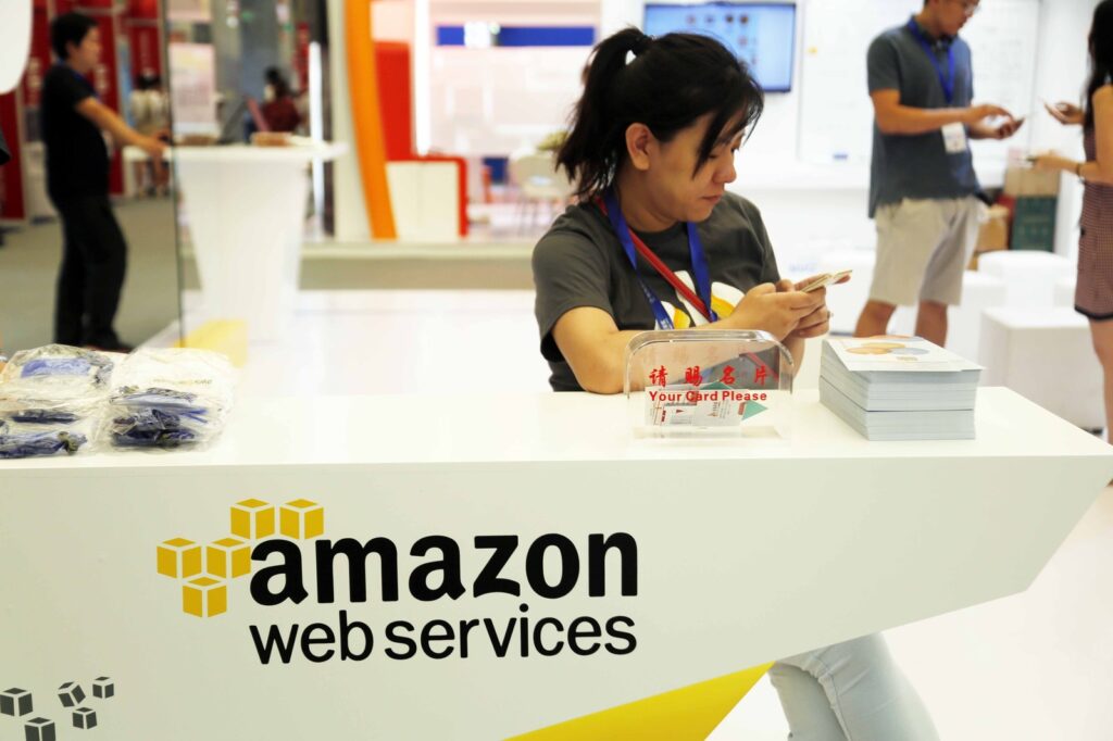 A woman sits at an Amazon Web Services booth, glancing at her phone amidst a sea of Amazon AWS promotional materials.