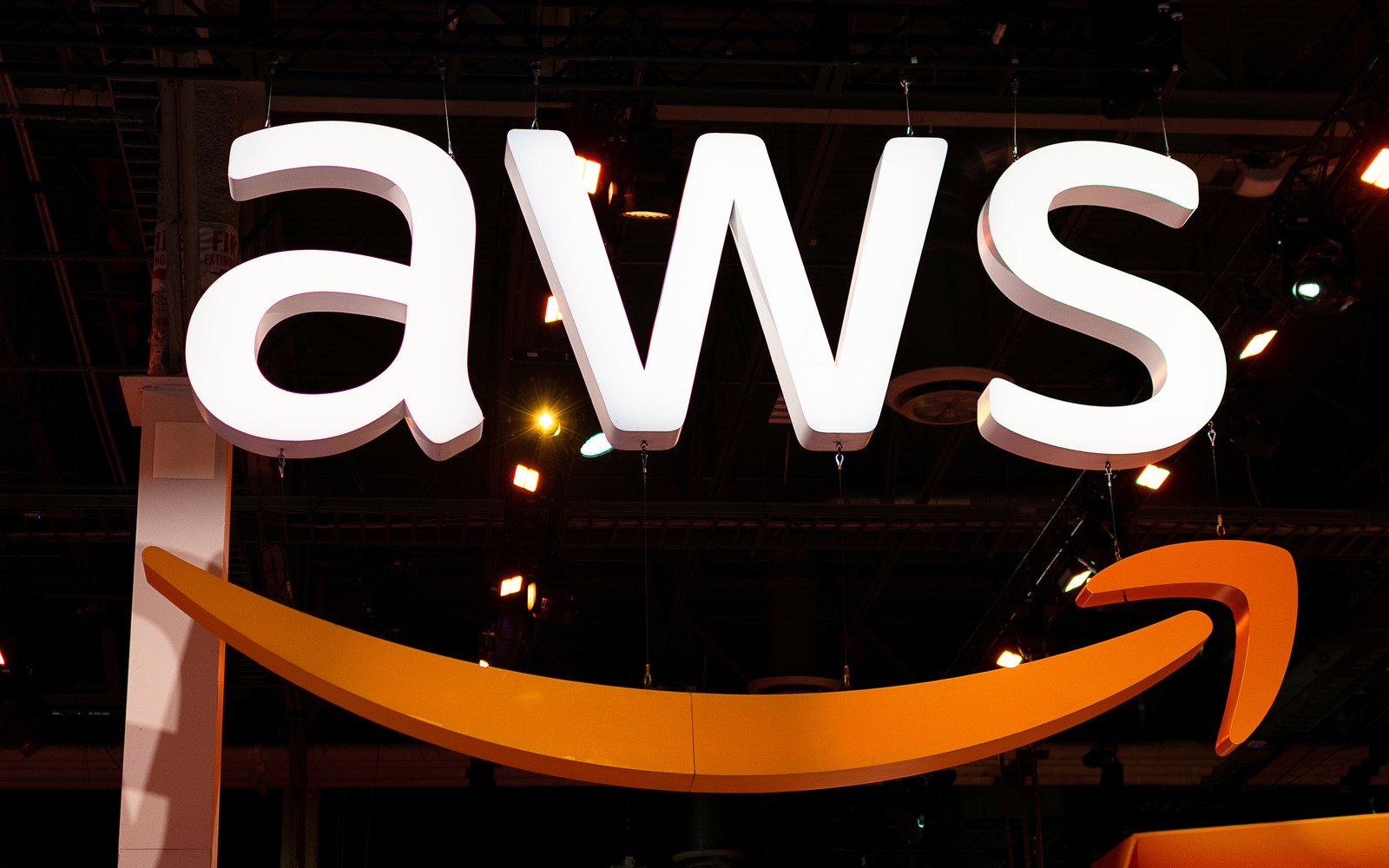 The Amazon AWS logo features large white letters AWS gracefully sitting above an orange curved arrow, all set against a dark background.